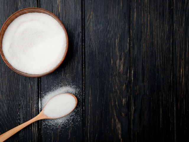 top view of white granulated sugar in a wooden bowl and in a wooden spoon on black rustic background with copy space