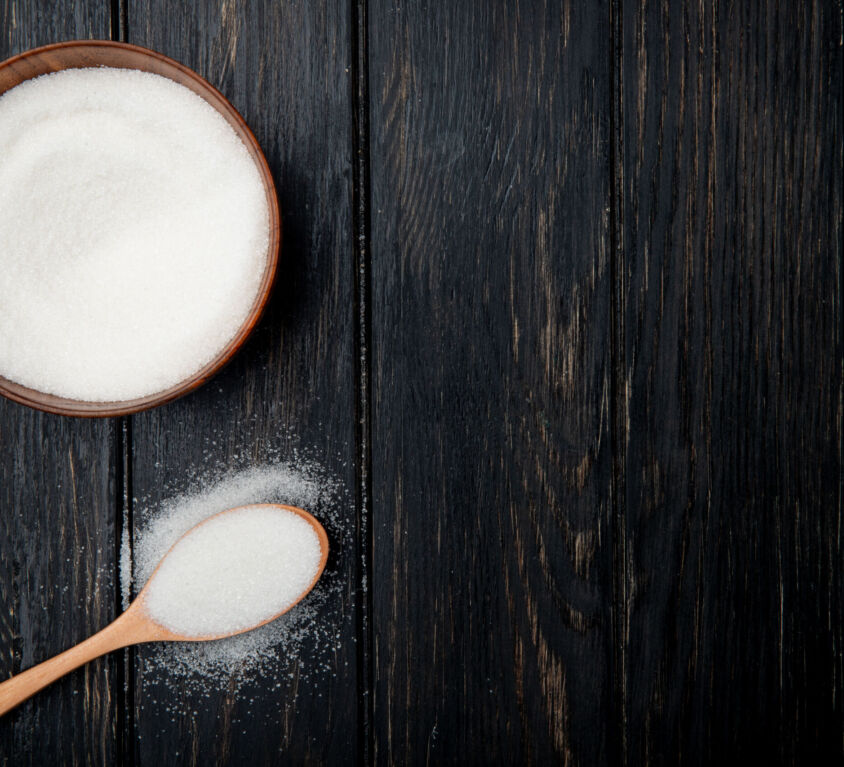 top view of white granulated sugar in a wooden bowl and in a wooden spoon on black rustic background with copy space