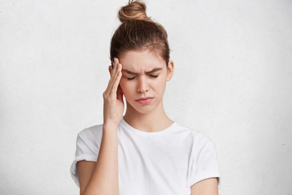 Portrait of tired exhausted young female has terrible headache after work, comes home in bad mood, being fed up with everything, needs rest, closes eyes and keeps hands on temples as feels pain