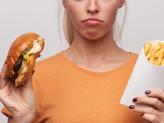 Upset young pretty blonde woman in orange t-shirt keeping unhealthy food in her hands and looking sadly at it, frowning eyebrows and twisting her mouth while posing over white background