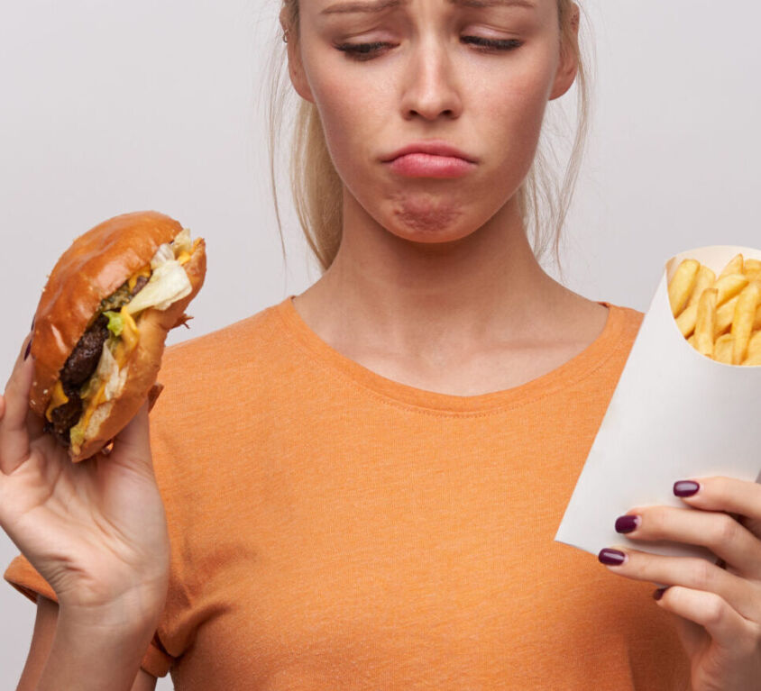 Upset young pretty blonde woman in orange t-shirt keeping unhealthy food in her hands and looking sadly at it, frowning eyebrows and twisting her mouth while posing over white background