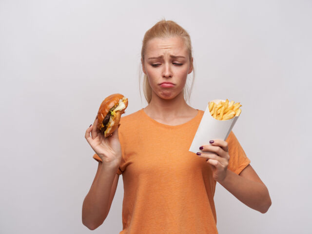 Upset young pretty blonde woman in orange t-shirt keeping unhealthy food in her hands and looking sadly at it, frowning eyebrows and twisting her mouth while posing over white background